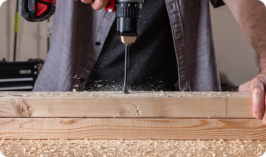 Cordless power drill boring into a wooden plank, with wood shavings scattered across the work surface