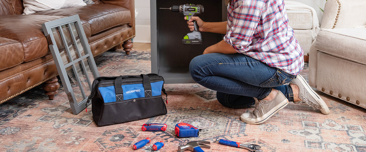 Repair worker using a cordless drill beside a metal vent cover, tool bag, and scattered hand tools on a rug