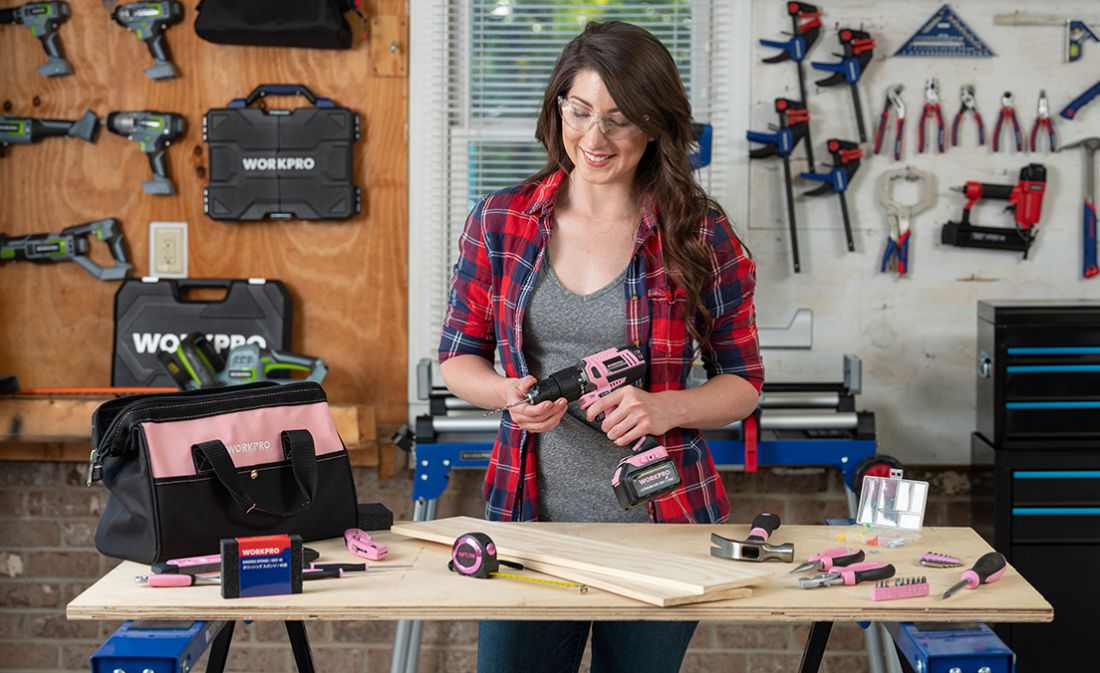 Woman holding a cordless drill in a workshop with tools, wood boards, a pink tool bag, and organized wall storage