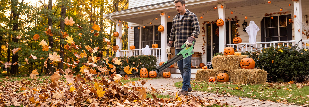 Man using a green leaf blower to clear fallen leaves outside a decorated house with pumpkins and hay bales