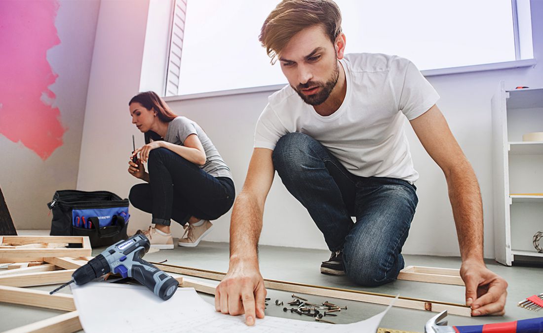 Couple installing wooden flooring with a cordless drill, hand tools, loose screws, and measuring materials nearby