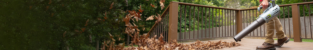 Person using a cordless leaf blower to clear dry leaves from a wooden deck beside a metal railing