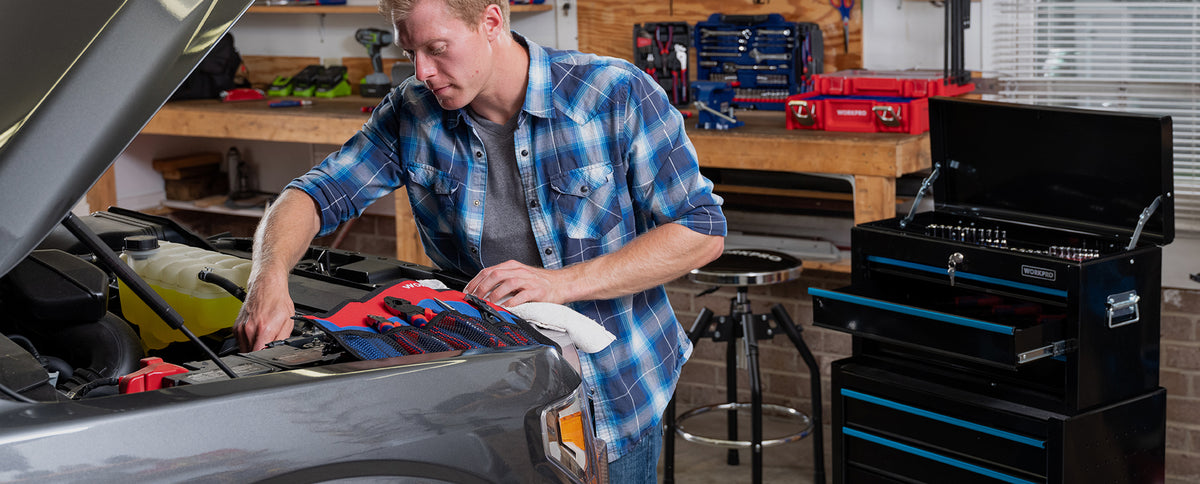 Mechanic using a red and blue tool set to repair a gray car beside a black rolling toolbox in a workshop