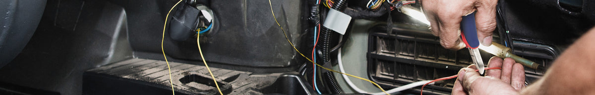 Hands repair exposed vehicle wiring beneath a dashboard with tools, cables, and a visible electrical connector