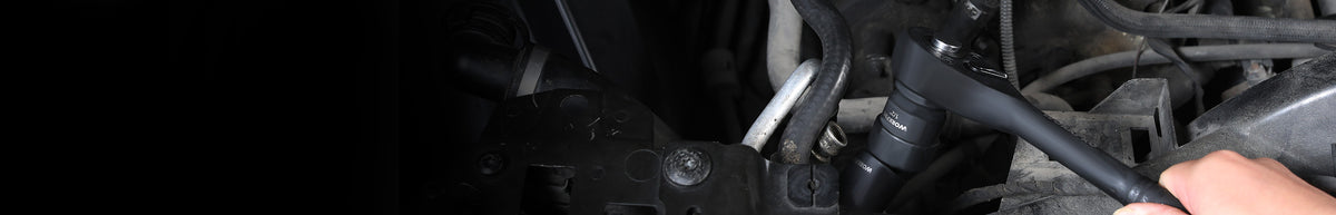 Close view of a vehicle engine compartment with black hoses, metal fittings, wiring, and a hand near the components